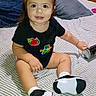 toddler, child, smiling, sitting, bed, striped_bedspread, black_onesie, hair_buns, indoor, can, white_socks, black_shoes, cute, person, furniture, pillow, wooden_cabinet, home, happy, young_child