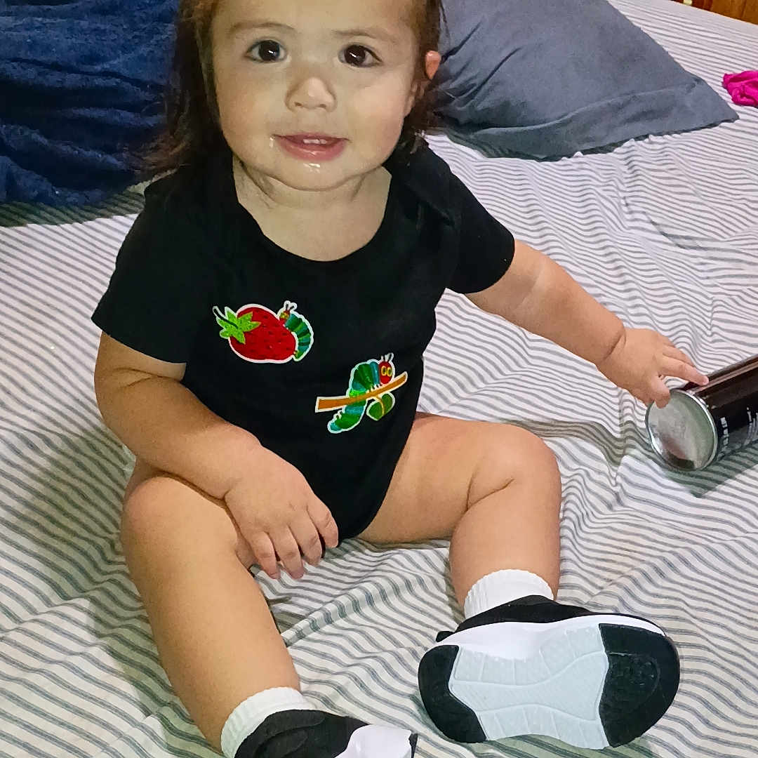 Alina is registered to the contest to win money with this photo: toddler, child, smiling, sitting, bed, striped_bedspread, black_onesie, hair_buns, indoor, can, white_socks, black_shoes, cute, person, furniture, pillow, wooden_cabinet, home, happy, young_child