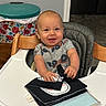 Tyler is registered to the contest to win money with this photo: baby, book, child, clothing, face, floor, floral_pattern, furniture, happy, high_chair, home, indoor, laundry_machine, person, play, room, smiling, table, trash_bin, wood
