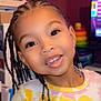 child, smiling, braids, indoor, colorful_clothing, flower_pattern, young_child, happy, portrait, face, person, toy, shelf, earrings, black_hair, pink, yellow, background, home, cute
