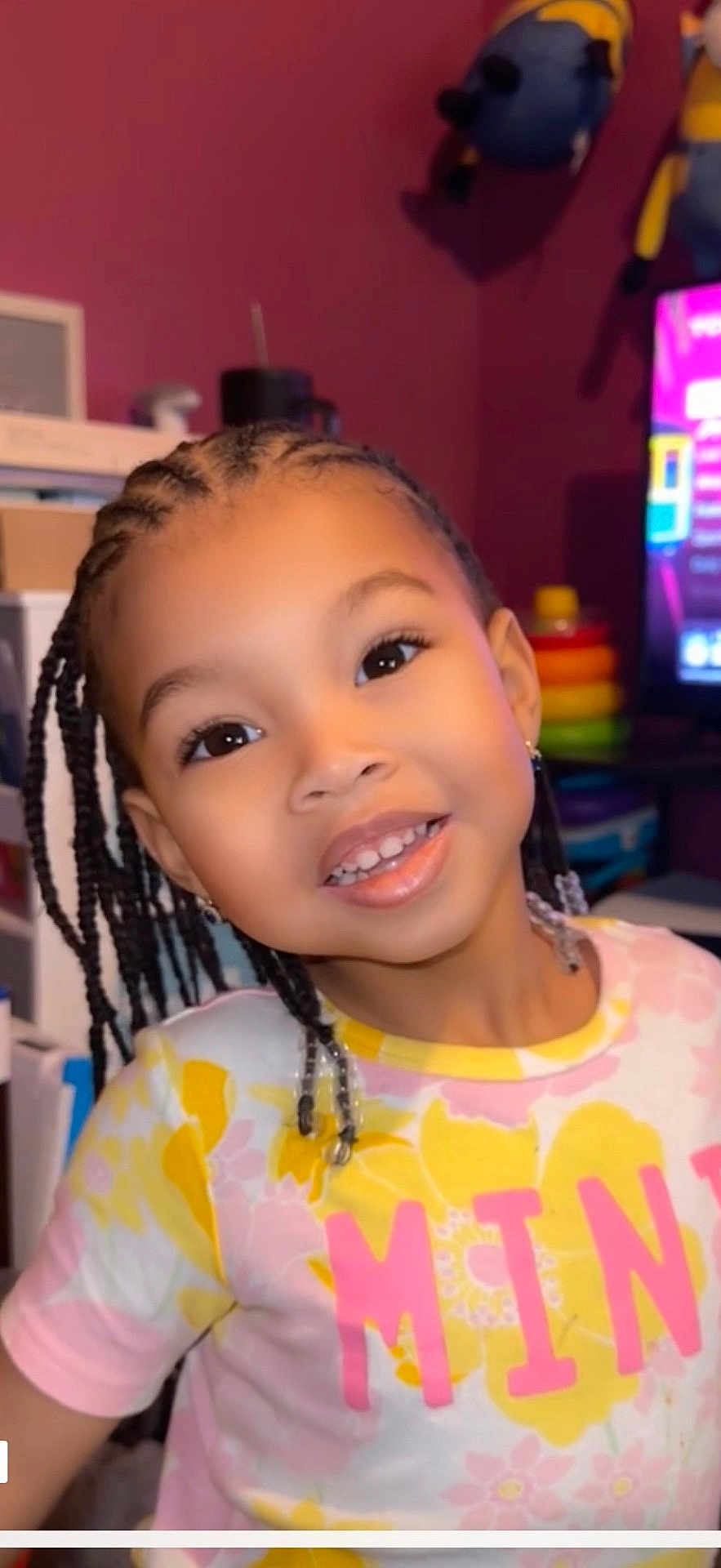 Nylah Arya joined the competition — help win amazing prizes! child, smiling, braids, indoor, colorful_clothing, flower_pattern, young_child, happy, portrait, face, person, toy, shelf, earrings, black_hair, pink, yellow, background, home, cute
