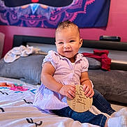 Nylah Arya joined the competition — help win amazing prizes! baby, bed, blanket, child, clothing, cute, decor, happy, home, indoor, infant, jeans, person, portrait, purple_shirt, sitting, smiling, socks, tapestry, wooden_sign