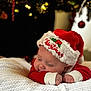 baby, christmas_hat, red_clothing, white_blanket, sleeping, holiday, festive, face, infant, cute, holiday_decor, christmas_tree, ornaments, soft_lighting, cozy, closeup, celebration, holidays, child, resting
