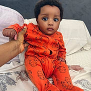 Zimuzochukwu joined the competition — help win amazing prizes! baby, infant, child, onesie, orange_clothing, bed, hand, person, cute, wide_eyes, animal_print, sitting, indoor, portrait, soft_lighting, calm_expression, bedroom, pillow, blanket, cozy