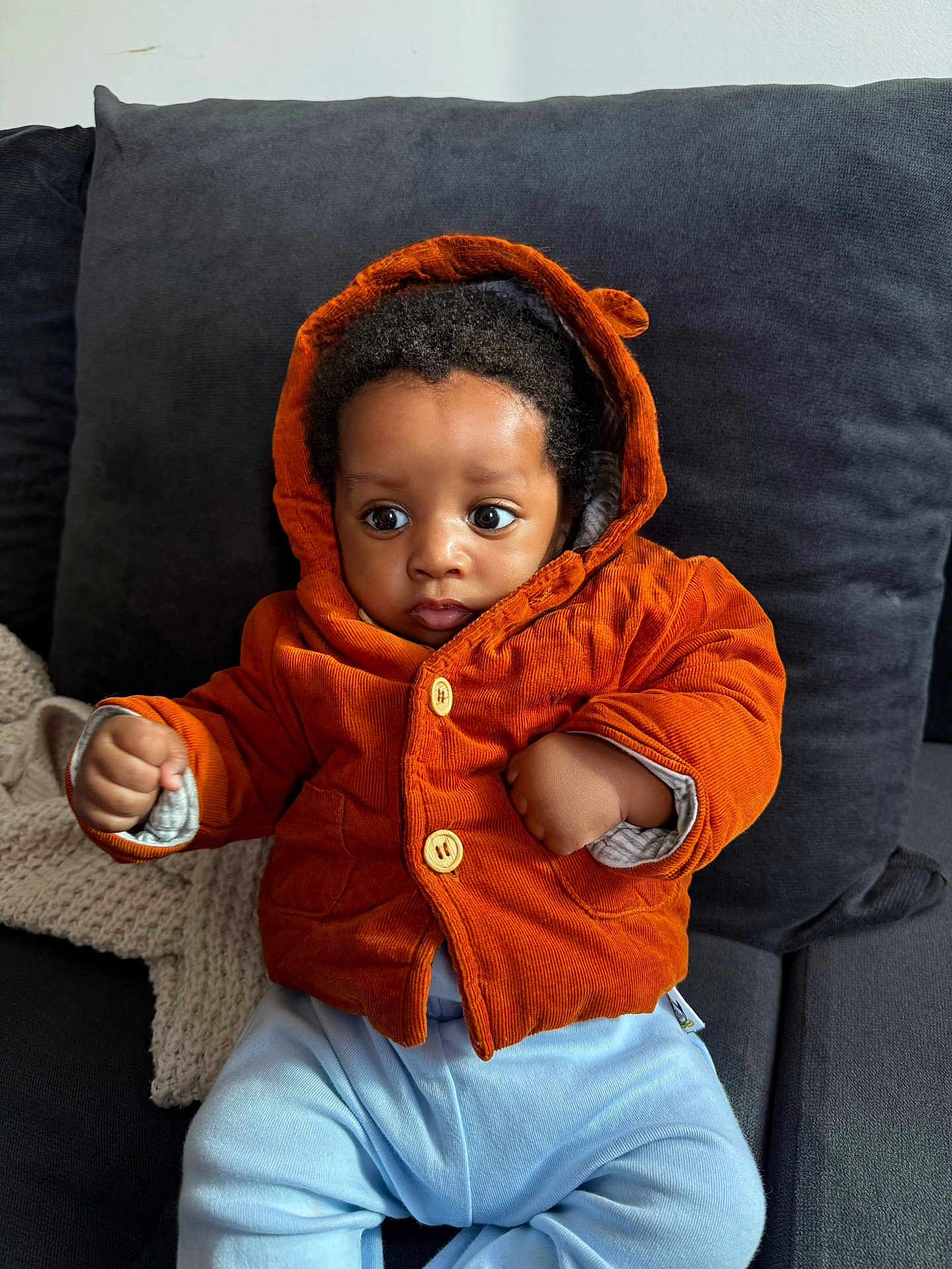 Zimuzochukwu joined the competition — help win amazing prizes! baby, child, hooded_jacket, orange_clothing, blue_pants, curly_hair, indoor, couch, pillow, knit_blanket, fist, wide_eyes, cute, portrait, seated, infant, warm_clothing, cozy, expression, person