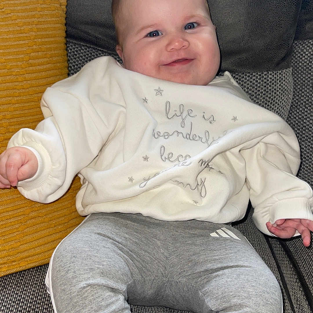 Elora joined the competition — help win amazing prizes! baby, casual_clothing, child, comfort, couch, cozy, face, gray_pants, happy, head, indoor, infant, pillow, portrait, relaxed, seated, smiling, sweatshirt, textile, white_socks