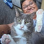 cat, person, glasses, paw, pink_pads, gray_cat, white_cat, feline, smile, couch, indoor, closeup, pet, happy, fur, whiskers, face, hand, tattoo, relaxed