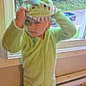 child, costume, dinosaur_costume, face, fashion, fuzzy, green, hat, home, hooded, indoors, onesie, outdoors, parked_vehicle, playful, portrait, smiling, standing, trees, window