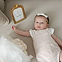 baby, infant, girl, romper, headband, bow, flower_bouquet, couch, pillow, gold_frame, sign, 2_months, soft_lighting, indoors, portrait, cute, newborn, lying_down, pastel_colors, decor