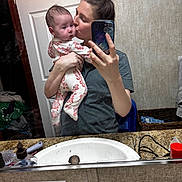 Eliza is registered to the contest to win money with this photo: baby, woman, mirror, selfie, bathroom, sink, door, clothing, holding, kissing, floral_pajamas, hair_bun, countertop, basket, towel, phone, reflection, person, indoor, portrait