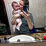 baby, woman, mirror, selfie, bathroom, sink, door, clothing, holding, kissing, floral_pajamas, hair_bun, countertop, basket, towel, phone, reflection, person, indoor, portrait