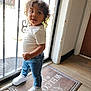 toddler, child, curly_hair, standing, white_shirt, jeans, white_sneakers, welcome_mat, door, glass_door, metal_bars, floor, wood_floor, indoor, natural_light, portrait, casual_clothing, home, smiling, looking_away