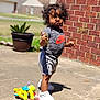 child, toddler, curly_hair, standing, toy, plush_turtle, concrete, brick_wall, potted_plant, outdoor, sunny, clothing, sneakers, shorts, camouflage, nike_logo, shadow, grass, house, sidewalk