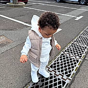 Warenn participe au concours pour gagner de l'argent avec cette photo : toddler, child, curious, white_clothing, vest, sneakers, parking_lot, metal_grate, asphalt, cars, tree, outdoor, sidewalk, street, urban, daytime, person, standing, looking_down, curly_hair