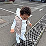 toddler, child, curious, white_clothing, vest, sneakers, parking_lot, metal_grate, asphalt, cars, tree, outdoor, sidewalk, street, urban, daytime, person, standing, looking_down, curly_hair