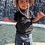 toddler, child, boy, hat, sun_hat, black_shirt, cargo_pants, water, fountain, outdoor, sunlight, shadow, smile, happy, standing, person, daylight, nature, playful, casual_clothing