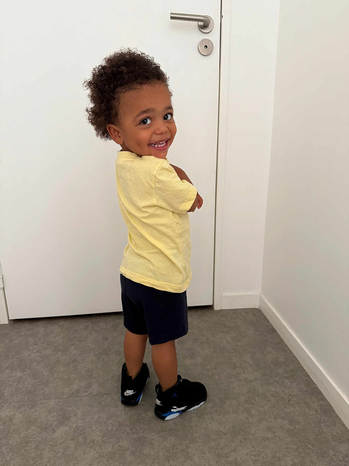 Warenn a rejoint le concours — aidez-le/la à gagner de superbes lots ! child, toddler, curly_hair, yellow_shirt, navy_shorts, black_sneakers, nike, indoor, door, floor, smiling, happy, playful, person, standing, looking_back, wall, casual_clothing, footwear, young