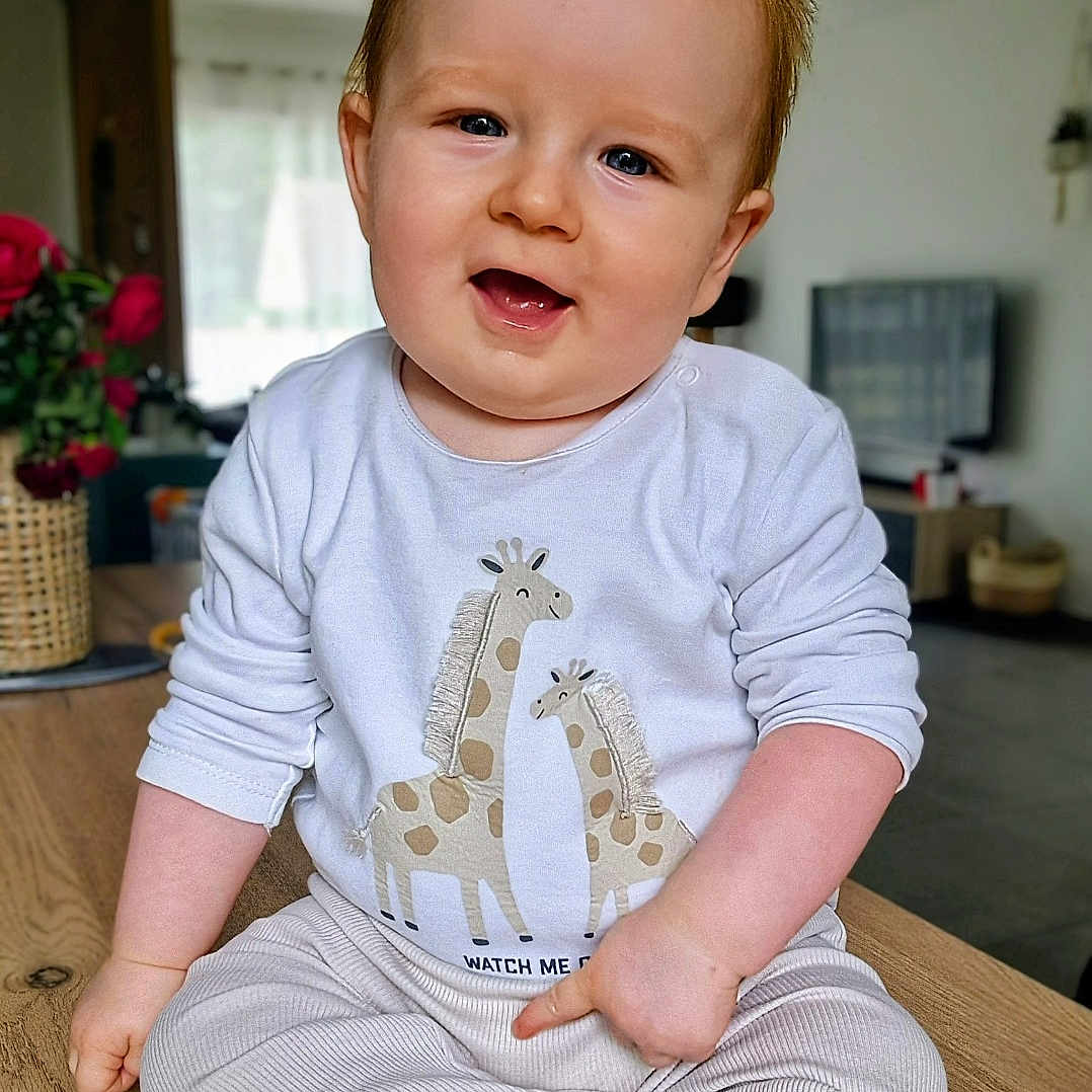 Alexis participe au concours pour gagner de l'argent avec cette photo : baby, barefoot, child, cozy, flowers, furniture, giraffe_print, happy, indoor, infant, living_room, long_sleeves, natural_light, pants, portrait, sitting, smiling, vase, white_shirt, wooden_table