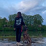 animal, calm, canine, cloudy_sky, dog, evening, footwear, interaction, landscape, leisure, mud, nature, outdoor, person, pet, reflection, river, sportswear, trees, water