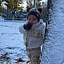 baby, beanie, cap, clothing, coat, face, footwear, glove, hat, head, jacket, knitwear, nature, outdoors, person, plant, snow, sweater, tree, treetrunk