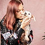 woman, dog, pet, kiss, leather_jacket, red_hair, small_dog, harness, tattoo, hand, pink_background, portrait, indoor, close_up, emotional, affection, casual_clothing, punk_style, sitting, human