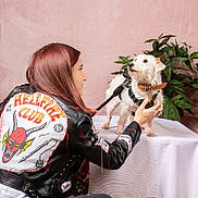Olympe participe au concours pour gagner de l'argent avec cette photo : person, dog, leather_jacket, table, plant, indoor, white_dog, pet, leash, collar, pink_background, green_plant, casual, portrait, hand, fashion, studio, cute, animal, interaction