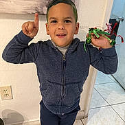 Dylan Galindo is registered to the contest to win money with this photo: child, boy, smiling, indoor, hoodie, jingle_bells, hand_raised, green_hair, tile_floor, wall, door, painting, casual_clothing, portrait, person, happy, young, playful, standing, home
