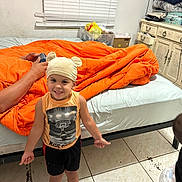 Dylan Galindo is registered to the contest to win money with this photo: barefoot, bear_ears, bed, bedroom, blanket, cabinet, casual, child, clothing, curtains, furniture, happy, hat, indoor, orange, person, smiling, tile_floor, toddler, window