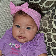 Amora is registered to the contest to win money with this photo: baby, infant, child, pink_headband, bow, purple_clothing, floral_pattern, couch, cushions, blue_eyes, portrait, close_up, cute, indoors, soft_lighting, curly_hair, smiling, face, person, young_child