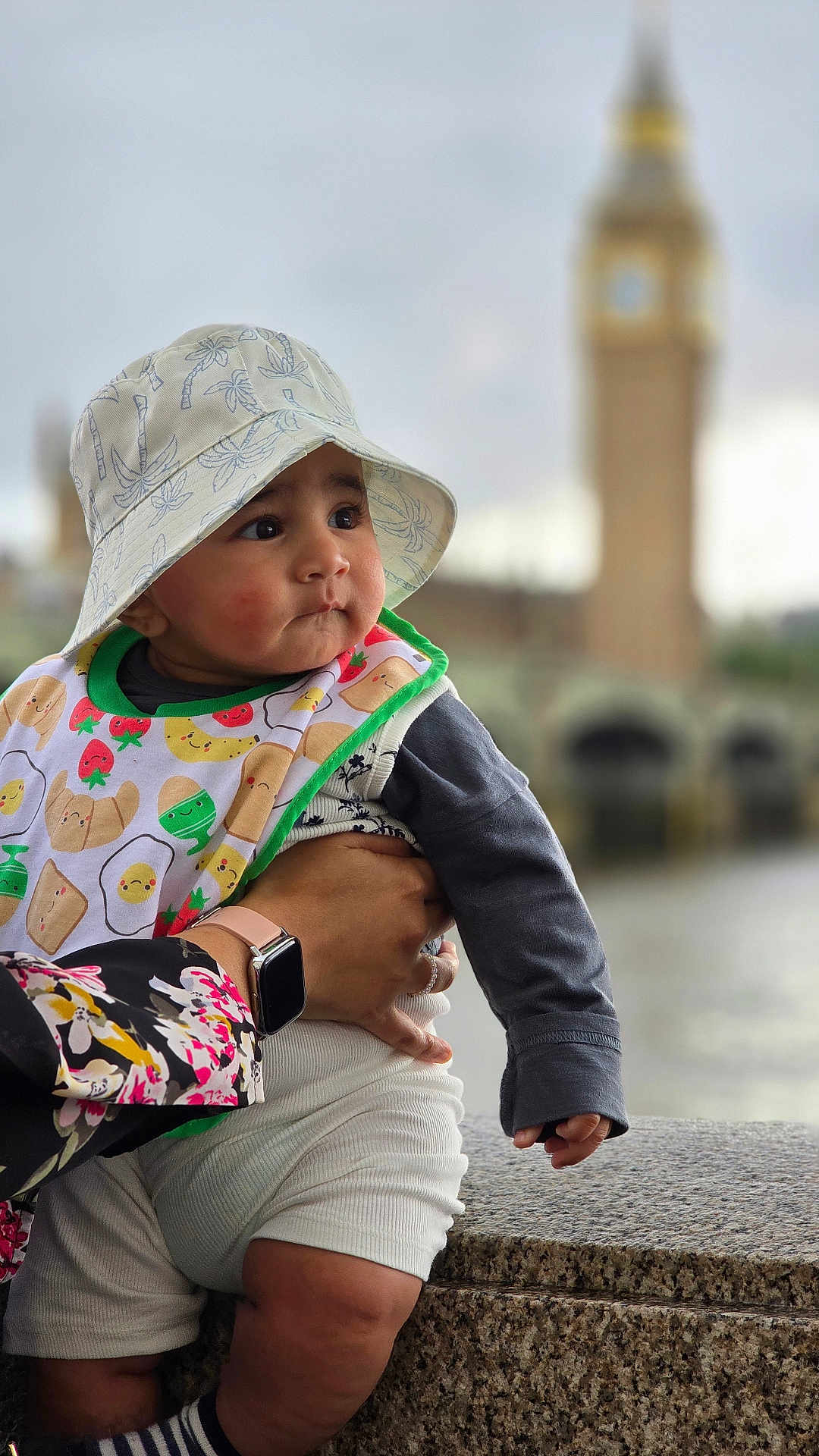 Rayaan is registered to the contest to win money with this photo: baby, child, hat, bib, hand, watch, sleeve, clock_tower, outdoor, portrait, candid, cute, smiling, colorful, shorts, striped_socks, granite_surface, blurred_background, person, holding