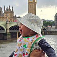 Rayaan joined the competition — help win amazing prizes! baby, child, bucket_hat, bib, river, big_ben, westminster_bridge, water, cityscape, person, happy, smiling, outdoor, landmark, clothing, hand, watch, sky, urban, tourist