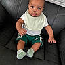baby, child, infant, sitting, bib, green_shorts, socks, black_couch, leather, curious, portrait, indoors, person, cute, young, small, looking, comfortable, clothing, expression