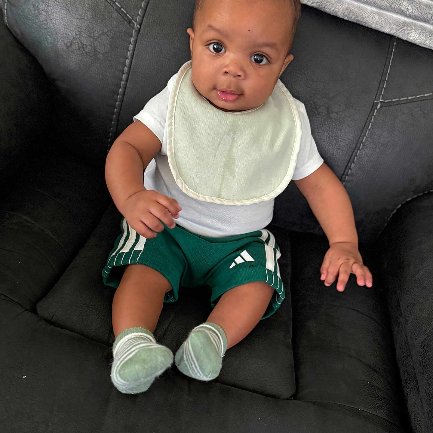 Zymeir joined the competition — help win amazing prizes! baby, child, infant, sitting, bib, green_shorts, socks, black_couch, leather, curious, portrait, indoors, person, cute, young, small, looking, comfortable, clothing, expression