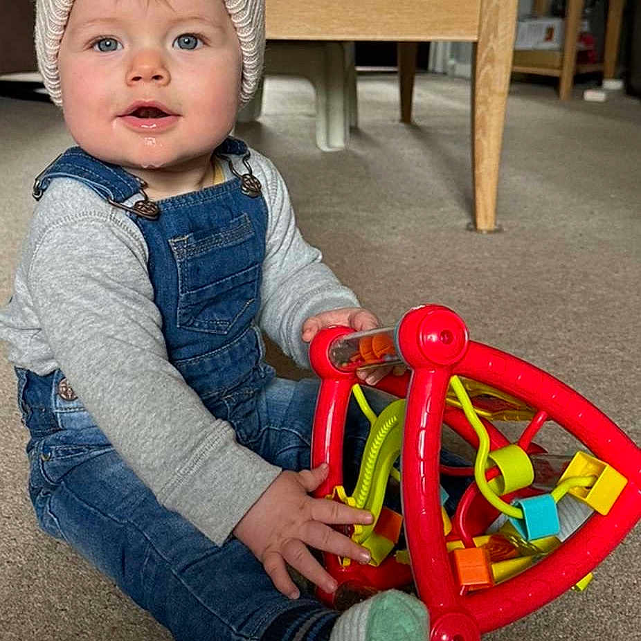 Ethan is registered to the contest to win money with this photo: baby, cap, clothing, couch, face, furniture, hardwood, hat, head, indoors, interiordesign, jeans, pants, person, photography, portrait, rattle, sitting, toy, wood
