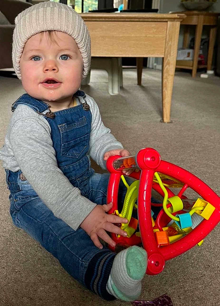 Ethan is registered to the contest to win money with this photo: baby, cap, clothing, couch, face, furniture, hardwood, hat, head, indoors, interiordesign, jeans, pants, person, photography, portrait, rattle, sitting, toy, wood