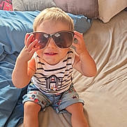 Evan a rejoint le concours — aidez-le/la à gagner de superbes lots ! baby, bed, casual_clothing, child, cute, fashion, footwear, happy, home, indoor, light, person, pillow, playful, shorts, sitting, smiling, striped_shirt, sunglasses, toddler