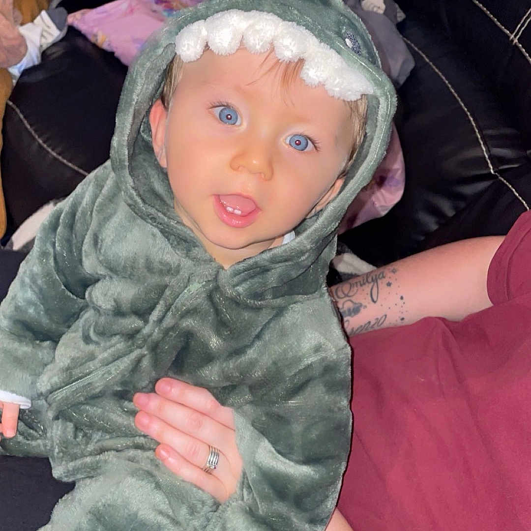 Emilio participe au concours pour gagner de l'argent avec cette photo : adult_hand, baby, black_leather, blue_eyes, child, couch, cozy, cute, dinosaur_costume, facial_expression, fuzzy_clothing, home, hood, indoor, onesie, person, plush, seated, smiling, tattoo
