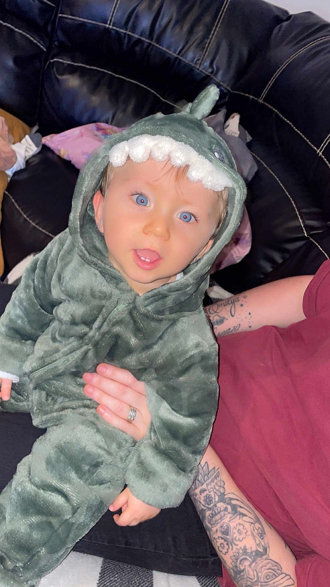 Emilio participe au concours pour gagner de l'argent avec cette photo : baby, child, dinosaur_costume, onesie, blue_eyes, couch, black_leather, tattoo, adult_hand, fuzzy_clothing, indoor, person, cute, smiling, seated, plush, hood, facial_expression, home, cozy