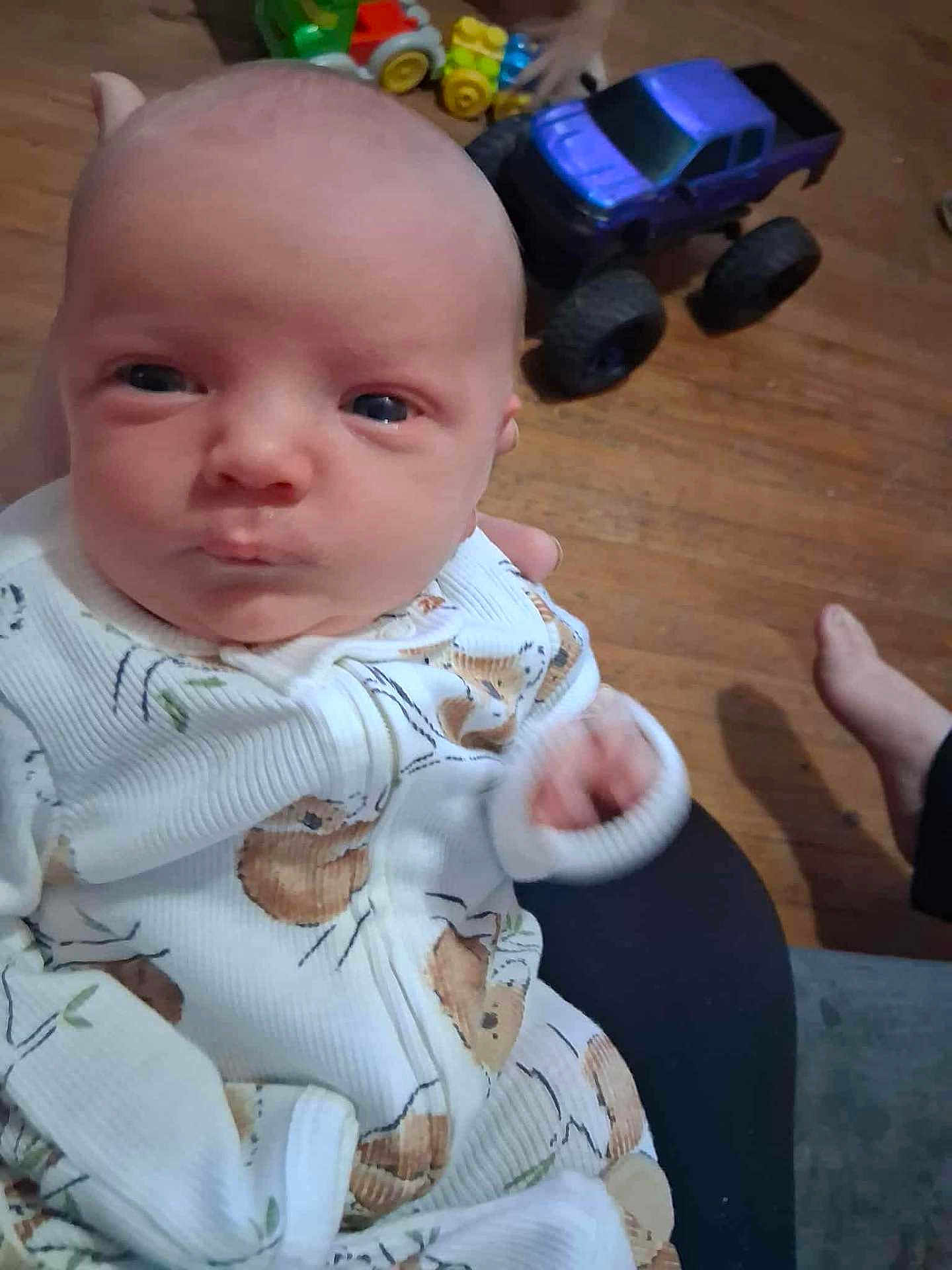 Aron is registered to the contest to win money with this photo: baby, infant, onesie, toy_truck, wooden_floor, hand, foot, person, indoor, child, clothing, plush_toy, furniture, flooring, couch, cute, portrait, closeup, human, baby_clothes