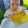 baby, child, high_chair, messy, food, yellow_food, face, hands, tongue_out, outdoor, grass, clothing, playful, happy, infant, cute, table, feeding, smiling, person
