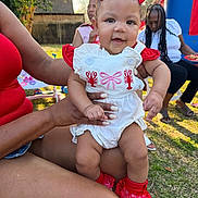 Daelanii is registered to the contest to win money with this photo: baby, child, red_bow, white_outfit, red_socks, white_shoes, adult_hand, outdoor, grass, inflatable, smiling, person, red_top, denim_shorts, sunny_day, woman, seated, tree, background, playground