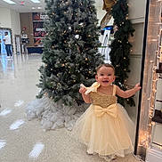 Amy is registered to the contest to win money with this photo: celebration, child, christmas_tree, decoration, dress, earrings, festive, floor, flooring, garland, gold, holiday_lights, indoor, joyful, person, pine_tree, smile, smiling, toddler, wall