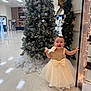 celebration, child, christmas_tree, decoration, dress, earrings, festive, floor, flooring, garland, gold, holiday_lights, indoor, joyful, person, pine_tree, smile, smiling, toddler, wall