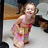 child, toddler, girl, pigtails, smiling, grinning, dress, colorful_clothing, standing, leaning, portrait, indoor, waiting_room, chairs, floor_tiles, wood_floor, exercise_equipment, background_person, casual_photo, playful