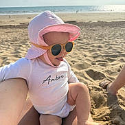 Ambre a rejoint le concours — aidez-le/la à gagner de superbes lots ! baby, child, sunglasses, pink_hat, beach, sand, sunhat, ocean, water, sunlight, person, legs, footwear, summer, outdoor, fun, playful, tongue_out, casual_clothing, vacation