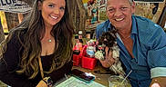 Tootsie Roll is registered to the contest to win money with this photo: adult, casual_clothing, dog, drinks, graffiti, leaves, man, menu, no_smoking_sign, outdoor, portrait, pub, puppy, restaurant, smiling, sunglasses, table, trees, water_bottle, woman