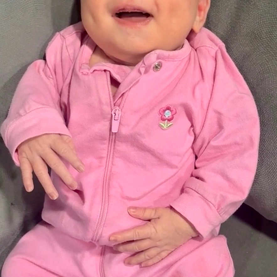 Natalie is registered to the contest to win money with this photo: baby, pink_clothing, smiling, happy, infant, couch, gray_furniture, indoor, portrait, child, one_person, seated, cute, cozy, relaxed, face, hands, footed_pajamas, zipper, flower_patch