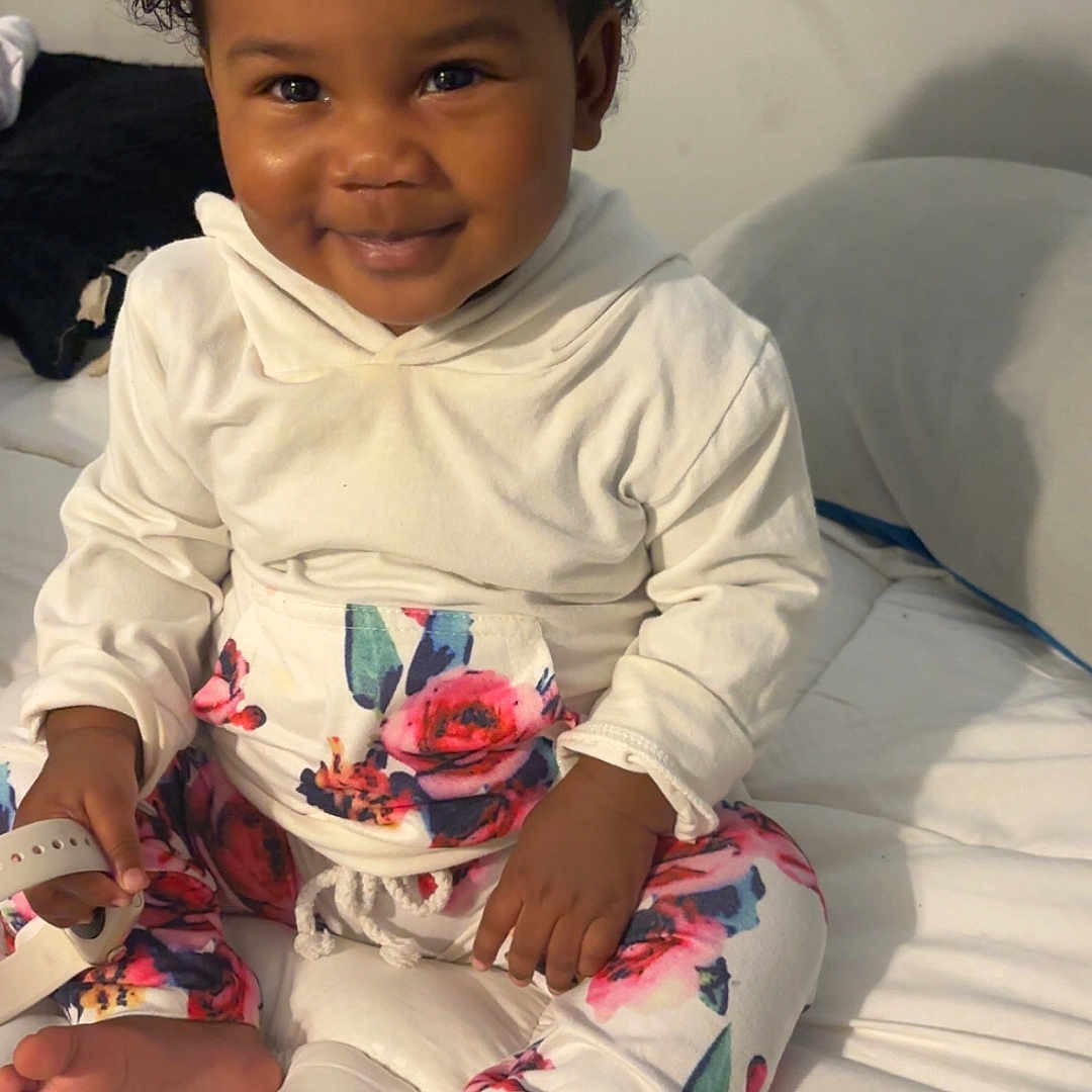 Amiyyah joined the competition — help win amazing prizes! barefoot, bed, blanket, child, cozy, curly_hair, cute, face, floral_pants, foot, hand, happy, indoors, person, sitting, skin, smiling, toddler, white_hoodie, young