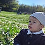 toddler, child, outdoor, grass, flowers, beanie, jacket, sunlight, nature, park, curious, sitting, daytime, greenery, portrait, young_child, casual_clothing, expression, person, scenic