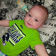 O'Ryen is registered to the contest to win money with this photo: baby, child, infant, smile, seahawks, green_shirt, bottle, bedspread, pattern, blue_eyes, cute, lying_down, plaid_pants, person, indoor, soft_lighting, happy, face, head, clothing